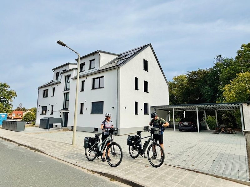 Visualisierung: zwei Radfahrer mit E-Bikes vor dem Haus, daneben der Carport